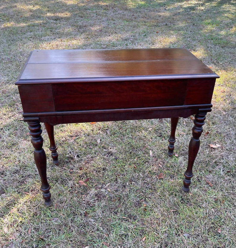 May include: A dark brown wooden writing desk with a drop-down front and turned legs. The desk is sitting on a green grassy surface.