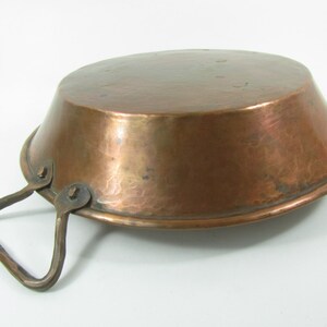 May include: A copper pan with a hammered finish and a single handle. The pan is empty and has a slightly flared rim.
