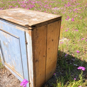 May include: A vintage wooden cabinet with a single door painted in a light blue color. The cabinet has a distressed finish and is sitting on a bed of pink flowers.