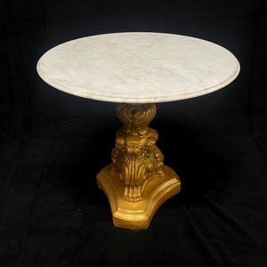 May include: A round white marble tabletop rests on a gold-colored pedestal base. The base features ornate, sculpted detailing with a flared, triangular foot. The surface of the marble shows subtle grey veining throughout the circular top.