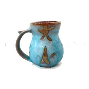 Puede incluir: Una taza pequeña de cerámica azul con un asa marrón y marcas marrones. La taza tiene un cuerpo redondeado y un borde ligeramente acampanado.