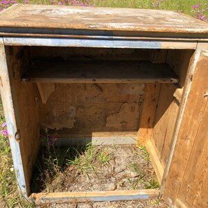 May include: A vintage wooden cabinet with a single door and a shelf inside. The cabinet is painted a light blue color with some of the paint chipped off. The cabinet is sitting on a grassy surface.