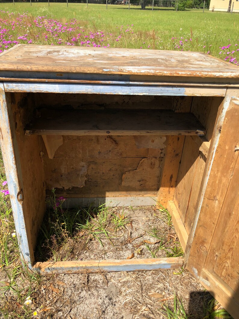 May include: A vintage wooden cabinet with a single door and a shelf inside. The cabinet is painted a light blue color with some of the paint chipped off. The cabinet is sitting on a grassy surface.