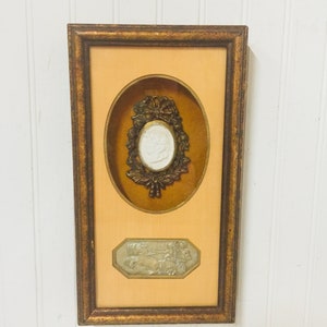 May include: A framed artwork with a gold-coloured frame and a cream-coloured mount. The artwork features a white cameo with a floral design and a smaller white cameo with a scene of people riding horses.