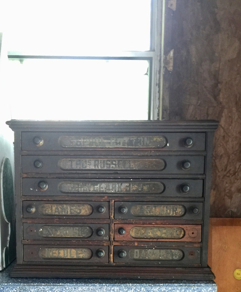 May include: Antique wooden drawer cabinet with eight drawers. Each drawer has a black label with white lettering. The labels read "S.C. COTTON", "THOMAS RUSSELL", "GEO. CLINTON", "JAMES", "GRIFFIN", "C.S. BULL", "NEVILLE", and "DAVIES".