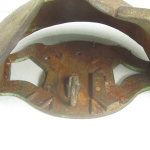 May include: A rusty metal object with a green patina. It has a circular base with a raised center and two hinged sections that open up. The object has a hook on the front and a series of holes around the edge.