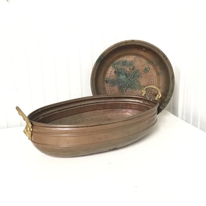 May include: An oval copper planter with two brass handles in the shape of lion heads. The planter has a matching copper lid with small holes for drainage.