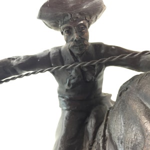 May include: A bronze sculpture of a cowboy riding a horse. The cowboy is wearing a hat and holding a rope. The sculpture is detailed and realistic.