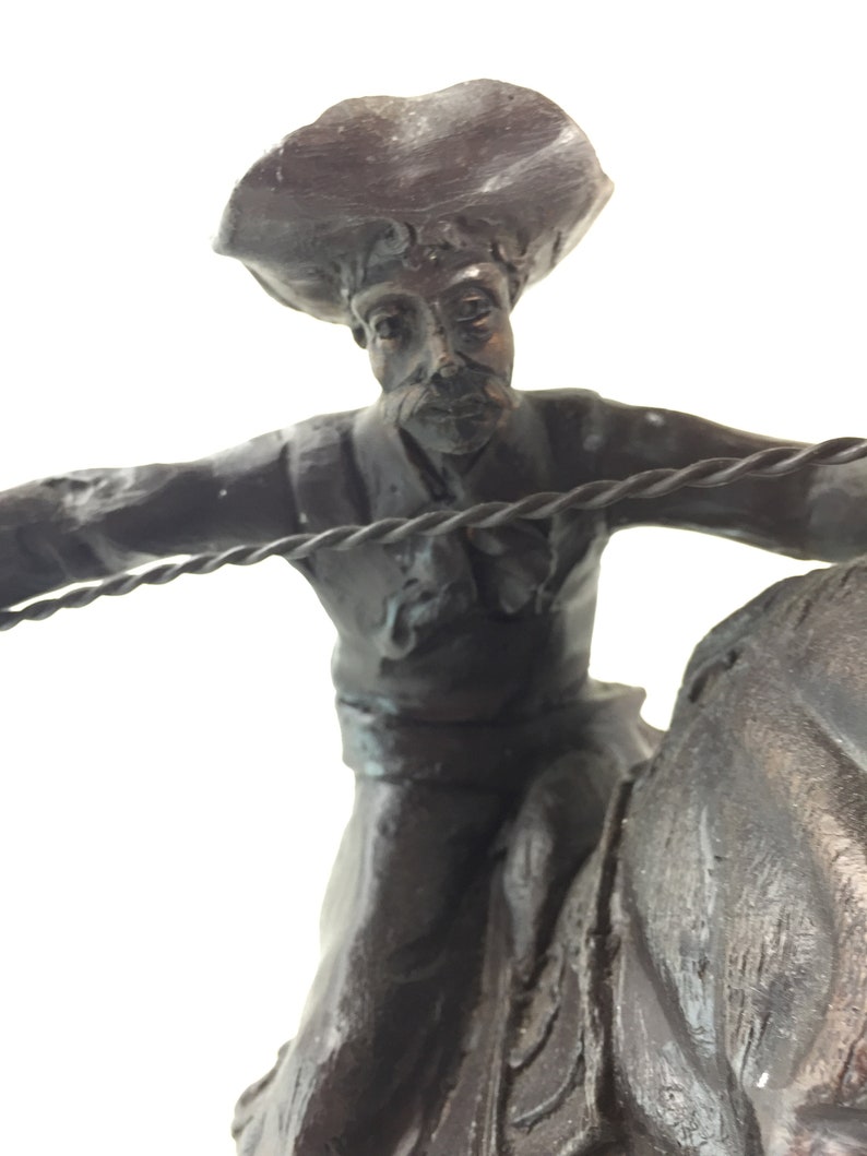 May include: A bronze sculpture of a cowboy riding a horse. The cowboy is wearing a hat and holding a rope. The sculpture is detailed and realistic.