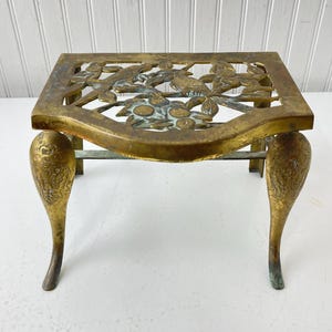May include: A vintage brass trivet with a rectangular top featuring a decorative openwork design of birds and flowers. The trivet has curved legs with ornate detailing. The brass has a warm, aged patina, adding to its antique charm.