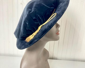 May include: A navy blue velvet hat with a gold tassel. The hat is displayed on a mannequin head. The hat has a soft, plush texture and a classic design. The gold tassel adds a touch of elegance.