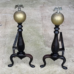 May include: A pair of black cast iron fireplace andirons with brass eagle finials and gold-colored ball tops. The andirons have a decorative, curved design.