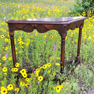 May include: A dark brown, antique wooden table with a decorative apron and four turned legs. The table is set in a field of yellow and pink wildflowers. The table's top is approximately 30 inches long.