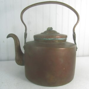 May include: A vintage copper kettle with a tarnished finish and a long, curved spout. The kettle has a single handle with a loop at the top.