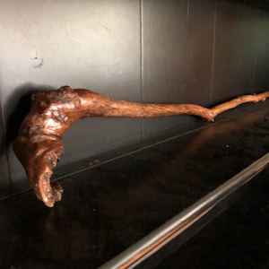 May include: A rustic wooden walking stick with a curved handle. The stick is brown and has a natural, weathered finish. It is resting on a black shelf.