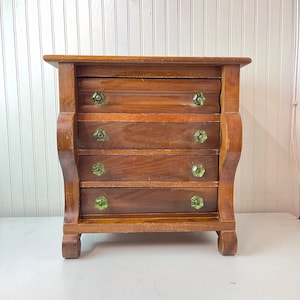 May include: A small wooden dresser with four drawers and green glass knobs. The dresser has a simple design with curved legs and a slightly rounded top.