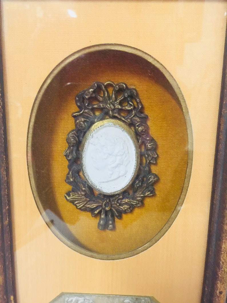 May include: A framed cameo with a white cameo set in a gold-toned metal frame with a decorative floral design.