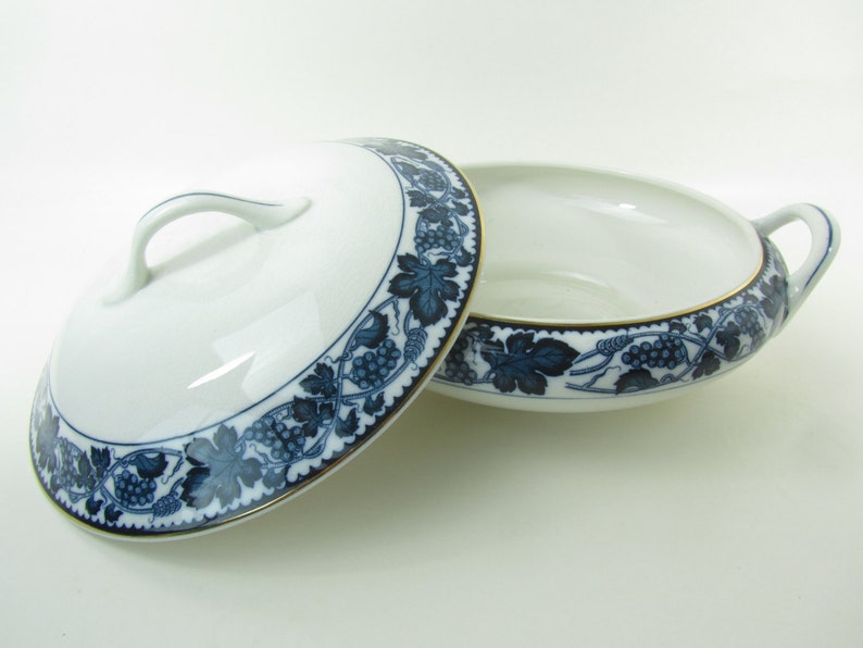 K&ouml;nnte beinhalten: Eine wei&szlig;e Porzellanservierschale mit einem blauen und goldenen Traubenmuster. Die Schale hat zwei Griffe und einen Deckel.