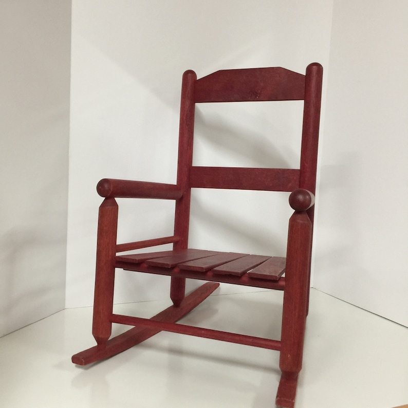 Puede incluir: Una silla mecedora de madera roja con un asiento de listones y brazos redondeados. La silla est&aacute; hecha de madera maciza y tiene un estilo r&uacute;stico, de granja.