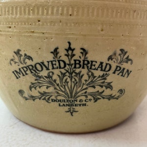 May include: Close-up of a vintage, off-white ceramic bread pan. The pan features the words "IMPROVED BREAD PAN" in black script, with decorative floral accents. The text "DOULTON & CO LAMBETH" is also visible. The pan has a textured surface.