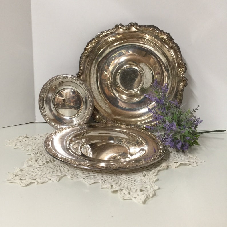 Puede incluir: Tres platos de servir plateados de diferentes tama&ntilde;os, con bordes ornamentados, dispuestos sobre un mantel de encaje blanco.