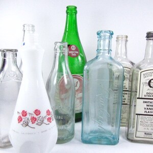 May include: Collection of vintage glass bottles, including a green glass bottle with a label that reads "Lemon-Lime Soda", a white glass bottle with a floral design, and a clear glass bottle with a label that reads "Watkins Imitation White Vanilla Flavor Double Strength".