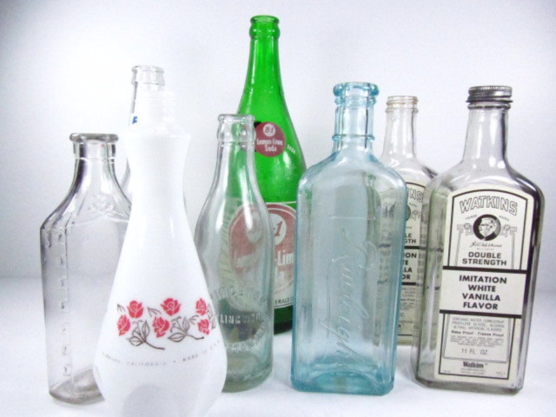 May include: Collection of vintage glass bottles, including a green glass bottle with a label that reads "Lemon-Lime Soda", a white glass bottle with a floral design, and a clear glass bottle with a label that reads "Watkins Imitation White Vanilla Flavor Double Strength".