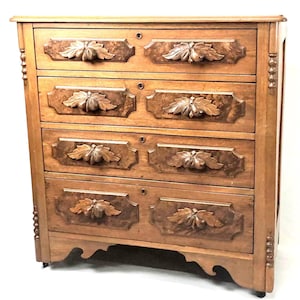 May include: A wooden dresser with four drawers. Each drawer has a decorative leaf design carved into the wood. The dresser is a light brown color and has a keyhole on each drawer. The sides of the dresser have decorative carvings.