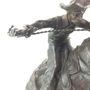 May include: A bronze sculpture of a cowboy riding a horse and throwing a lasso. The cowboy is wearing a hat and has a determined expression on his face. The horse is rearing up on its hind legs.