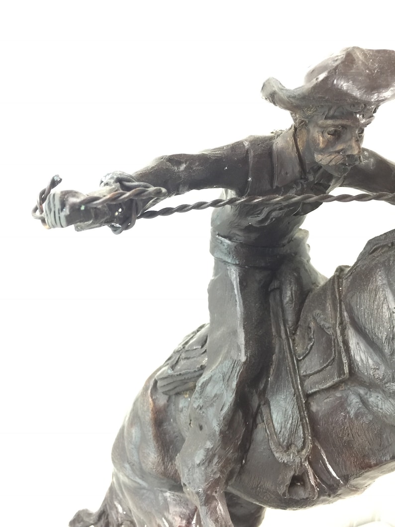May include: A bronze sculpture of a cowboy riding a horse and throwing a lasso. The cowboy is wearing a hat and has a determined expression on his face. The horse is rearing up on its hind legs.