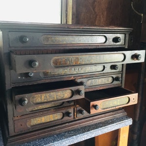 May include: Antique wooden drawer cabinet with multiple drawers, each labeled with a different name. The drawers are partially open, revealing the interior of the cabinet.