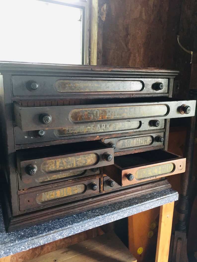 May include: Antique wooden drawer cabinet with multiple drawers, each labeled with a different name. The drawers are partially open, revealing the interior of the cabinet.