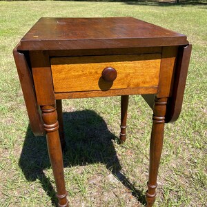 May include: A small, antique wooden table with a drawer and drop leaves. The table is made of dark brown wood with a lighter-colored drawer front and a round wooden knob. The table has four turned legs and is set on a grassy surface.