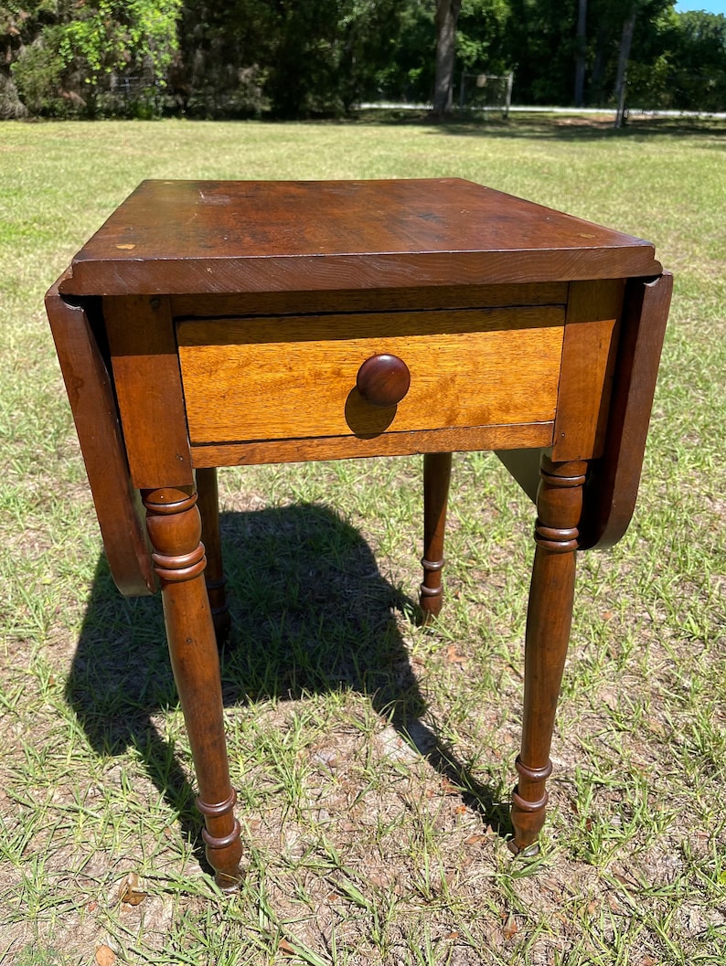 May include: A small, antique wooden table with a drawer and drop leaves. The table is made of dark brown wood with a lighter-colored drawer front and a round wooden knob. The table has four turned legs and is set on a grassy surface.