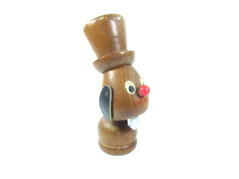 May include: A wooden figurine of a rabbit wearing a top hat. The rabbit has a red nose, black ears, and a white mouth. The figurine is brown with painted details.
