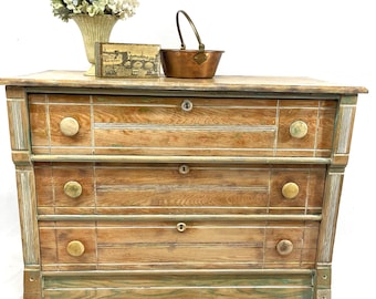 May include: A light-colored wooden dresser with three drawers and round knobs. The dresser has a distressed finish with green accents. A small copper pot, a decorative box, and a vase of flowers sit on top.