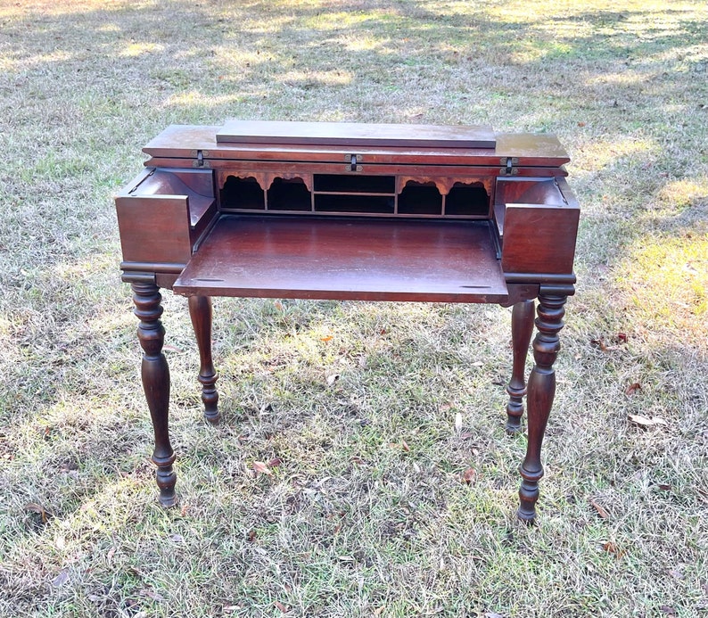 May include: A dark wood antique writing desk with a drop-down front and a series of small drawers. The desk has turned legs and is sitting on a green grassy surface.