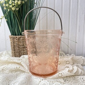 May include: A pink glass ice bucket with a silver handle. The bucket features a decorative etched design and is resting on a white lace tablecloth.