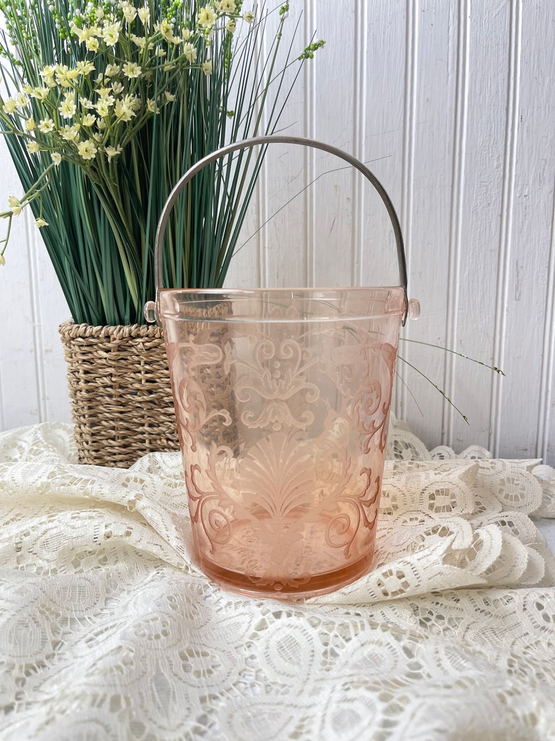 May include: A pink glass ice bucket with a silver handle. The bucket features a decorative etched design and is resting on a white lace tablecloth.