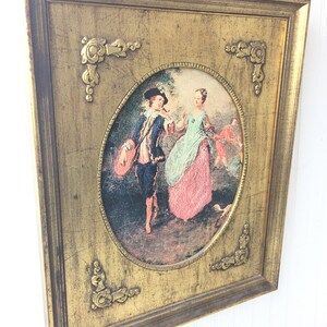 May include: A framed oval print of a painting depicting a couple in a garden setting. The man is wearing a blue suit and a hat, and the woman is wearing a pink dress. The frame is gold with ornate details.