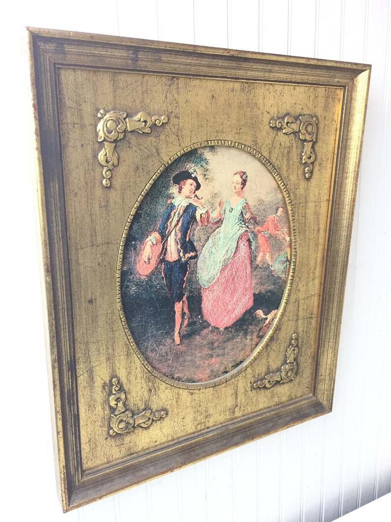 May include: A framed oval print of a painting depicting a couple in a garden setting. The man is wearing a blue suit and a hat, and the woman is wearing a pink dress. The frame is gold with ornate details.