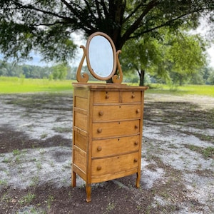Puede incluir: Una c&oacute;moda de madera vintage con un frente curvo y cinco cajones. La c&oacute;moda tiene un peque&ntilde;o espejo ovalado en la parte superior.