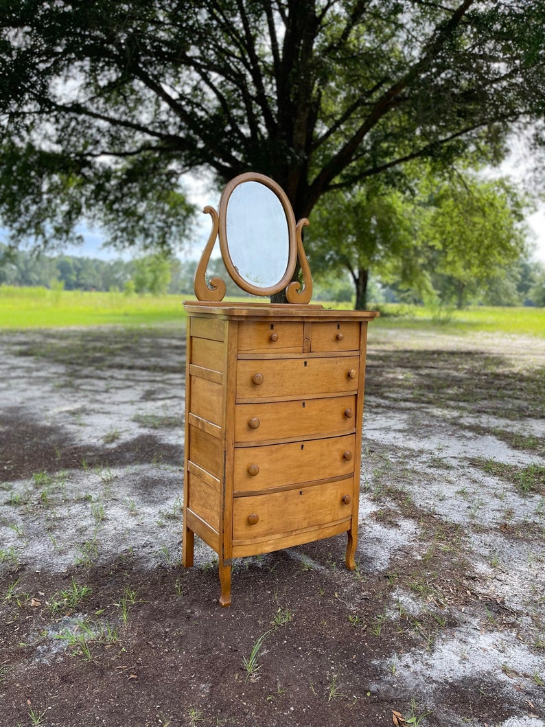 Puede incluir: Una c&oacute;moda de madera vintage con un frente curvo y cinco cajones. La c&oacute;moda tiene un peque&ntilde;o espejo ovalado en la parte superior.