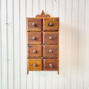 May include: A wooden spice rack with eight small drawers, each with a knob. The rack is mounted on a white wall.