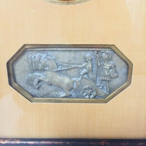 May include: A framed artwork featuring a silver-toned relief sculpture of a chariot pulled by a horse with two figures in the chariot. The artwork is framed in a brown wood frame with a gold-coloured inner border.