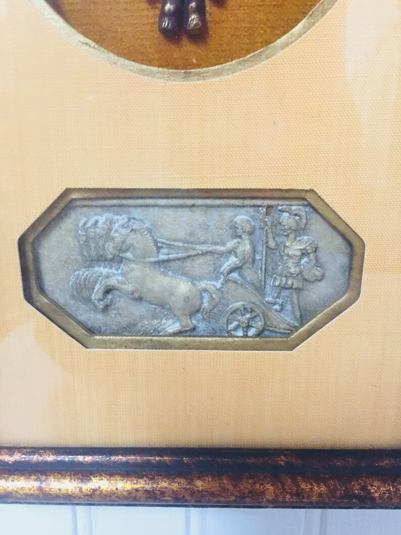 May include: A framed artwork featuring a silver-toned relief sculpture of a chariot pulled by a horse with two figures in the chariot. The artwork is framed in a brown wood frame with a gold-coloured inner border.