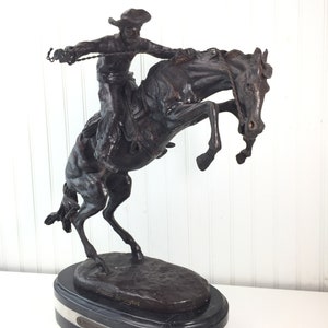 May include: A bronze sculpture of a cowboy riding a bucking horse. The sculpture is on a black marble base with a white marble band. The sculpture is signed "Frederic Remington" and "The Bronco Buster".