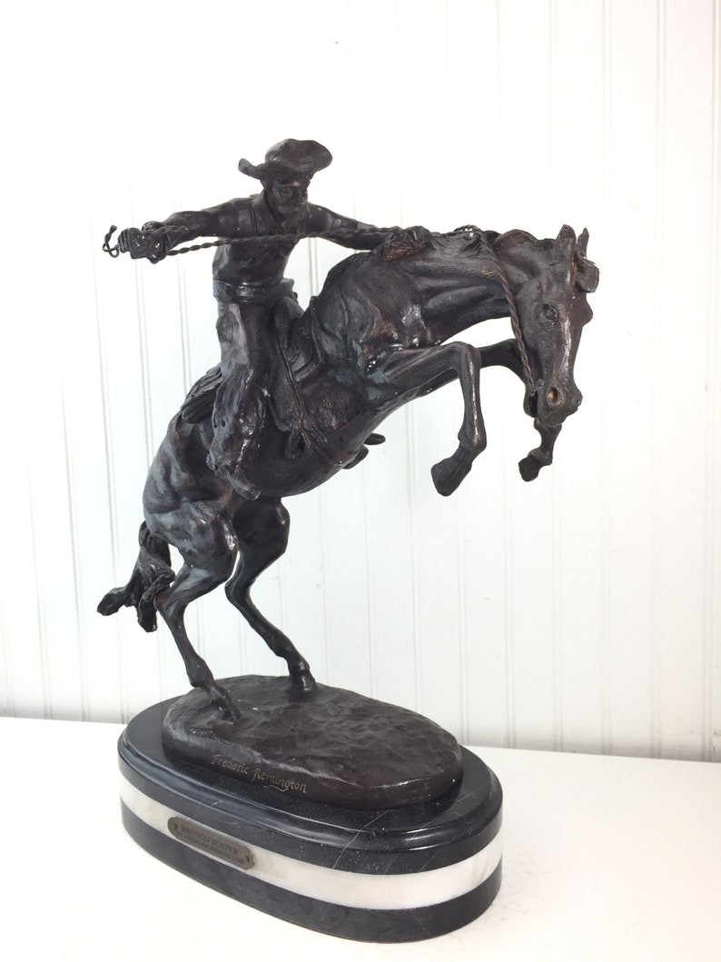 May include: A bronze sculpture of a cowboy riding a bucking horse. The sculpture is on a black marble base with a white marble band. The sculpture is signed "Frederic Remington" and "The Bronco Buster".