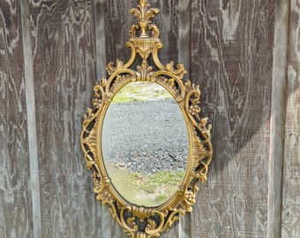 May include: A gold-toned oval mirror with an ornate, carved frame. The mirror is hanging on a wooden wall.