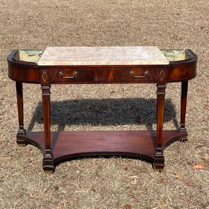 May include: A mahogany console table with a marble top and a single drawer. The table has a curved front and a lower shelf. The table is in a traditional style and has brass hardware.